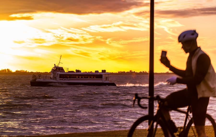 Ciclista parado na orla fotografando o barco que passa, com o céu dourado ao fundo