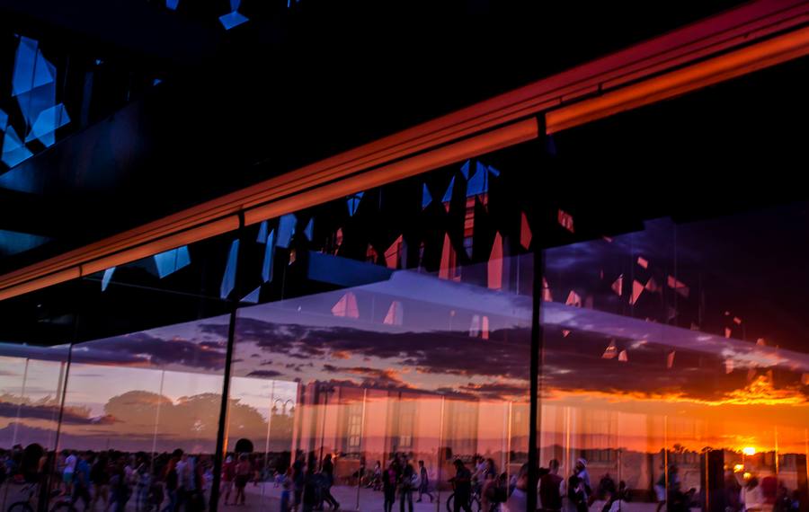 Bar na orla com o pôr do sol refletido nos vidros e pessoas à mesa