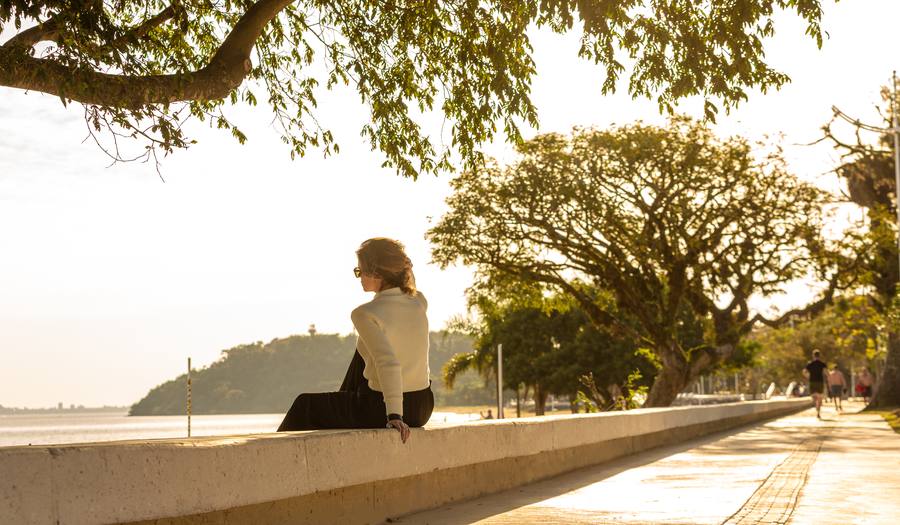 Mulher sentada no muro da orla, sob as árvores, olhando a água